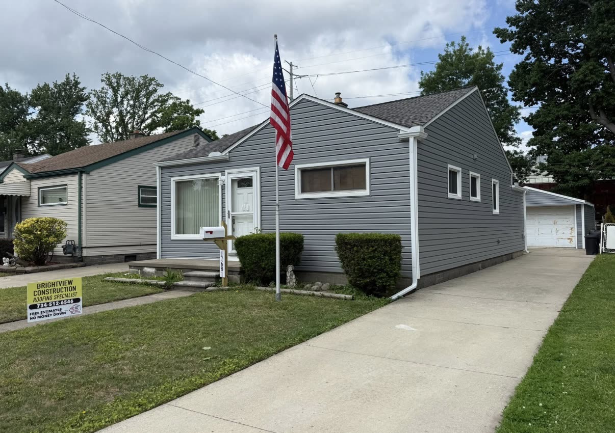 Completed dark asphalt shingle roof on a grey sided home with a BrightView Construction yard sign and an American flag in the front yard