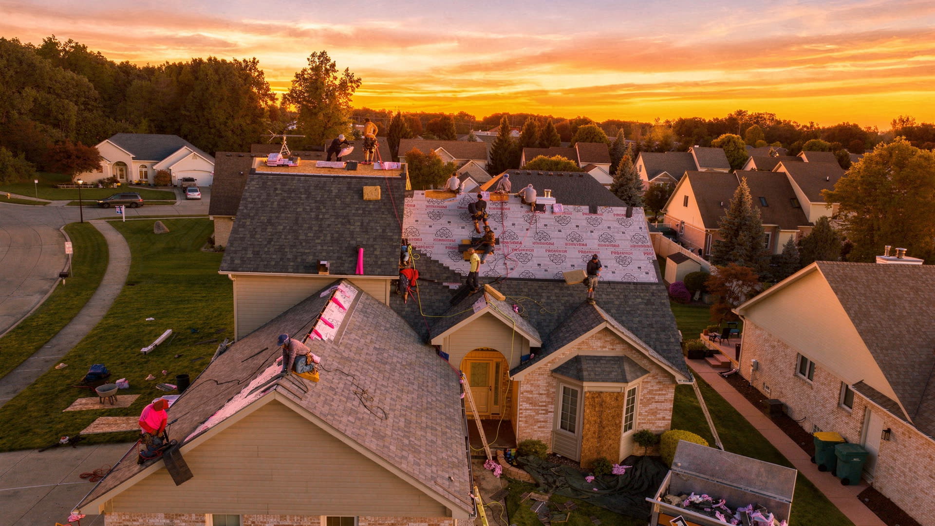 Aerial view at dusk of a BrightView Construction crew installing synthetic underlayment and stacking shingle bundles on a two story home